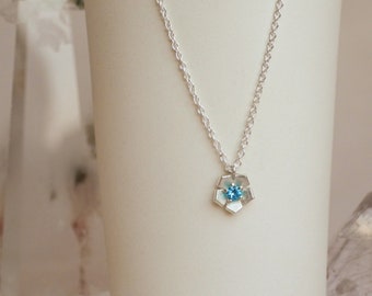 Pode incluir: Um colar de prata com um pingente em forma de flor. O pingente tem uma pedra preciosa azul clara no centro e um design geométrico. O colar é exibido em uma superfície branca, com formações de cristal ao fundo.