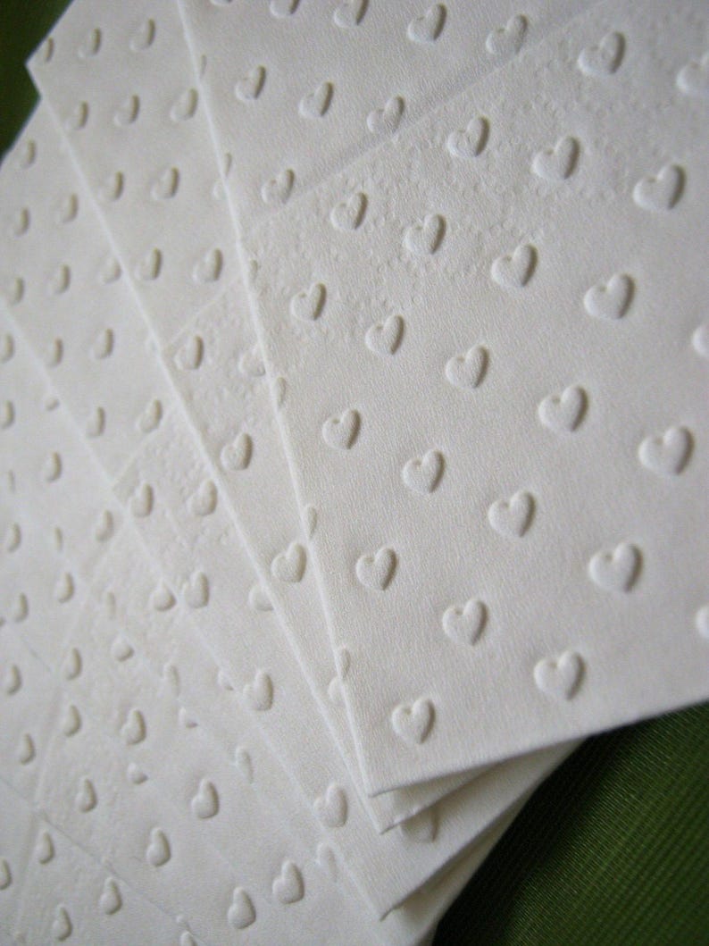 Puede incluir: Un paquete de servilletas de papel blancas con un sutil patr&oacute;n de corazones en relieve. Las servilletas est&aacute;n dobladas en forma de abanico.