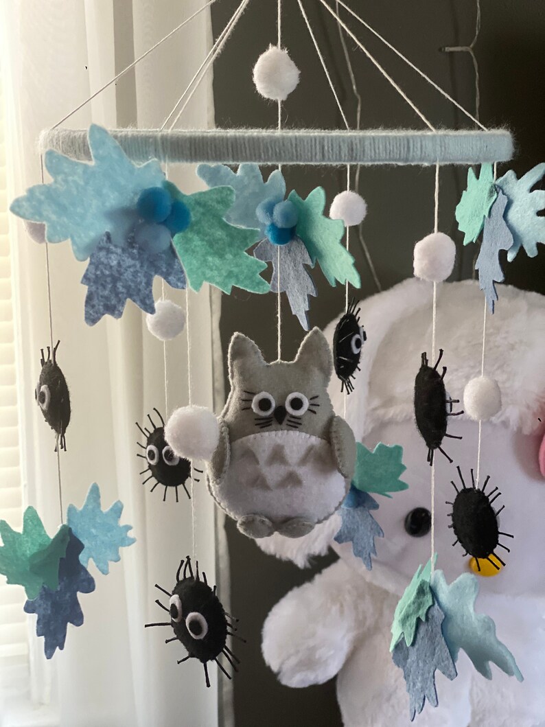 May include: A handmade baby mobile featuring felt leaves in shades of blue and green, white pom-poms, and a gray Totoro character. Black soot sprites dangle below. The mobile is suspended from a light blue ring.
