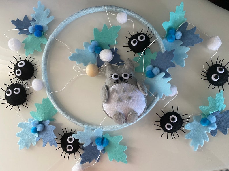 May include: A handmade mobile featuring a light blue ring adorned with felt leaves in shades of blue and white pom-poms. A gray Totoro figure and black soot sprites are attached, creating a whimsical nursery decoration.