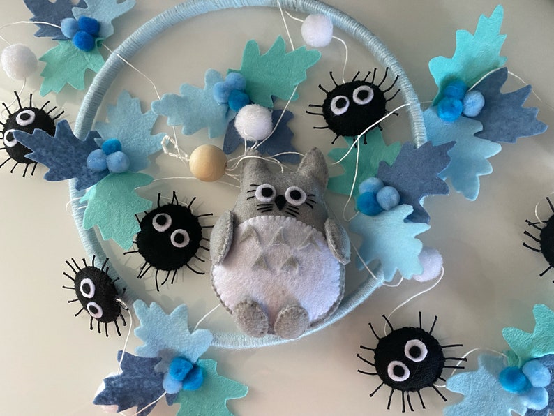 May include: A handmade felt garland featuring a gray Totoro character, black soot sprites, and blue leaf accents. The garland is strung on a white string with blue pom-poms and wooden beads, perfect for nursery decor.