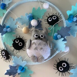 May include: A handmade dream catcher featuring a gray Totoro character, black soot sprites, and blue felt leaves. The design includes white pom-poms and wooden beads, all set against a light blue hoop.
