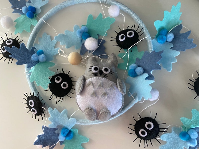 May include: A handmade dream catcher featuring a gray Totoro character, black soot sprites, and blue felt leaves. The design includes white pom-poms and wooden beads, all set against a light blue hoop.