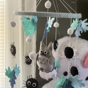 May include: A handmade baby mobile featuring felt characters and shapes. The mobile includes a gray Totoro figure, black soot sprites, and blue and green leaf shapes. White pom-poms and a light blue ring complete the design, suspended by white strings.