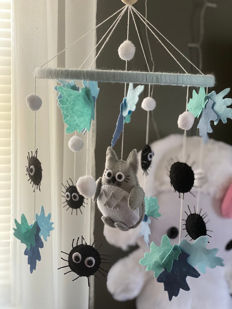 May include: A handmade baby mobile featuring felt characters and shapes. The mobile includes a gray Totoro figure, black soot sprites, and blue and green leaf shapes. White pom-poms and a light blue ring complete the design, suspended by white strings.