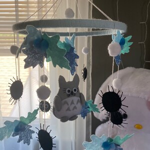 May include: A handmade baby mobile featuring a light blue ring wrapped in yarn, with felt leaves, pom-poms, and a gray Totoro character. The mobile also includes black, spiky soot sprites, creating a whimsical nursery decoration.