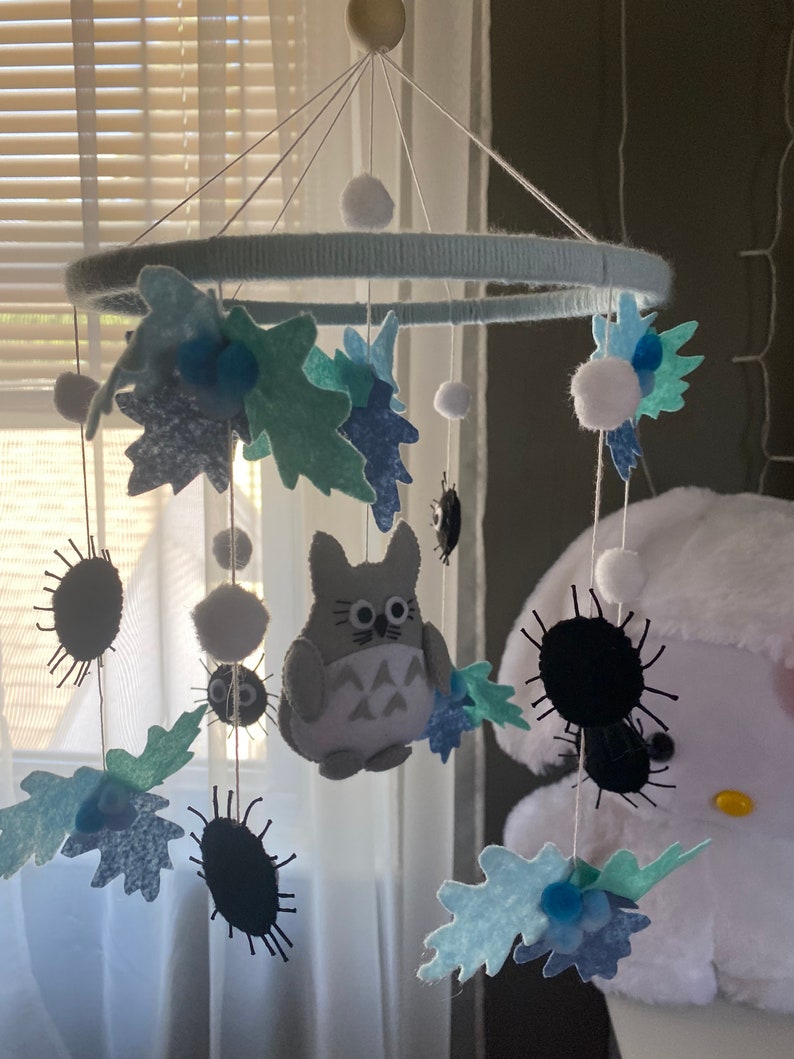 May include: A handmade baby mobile featuring a light blue ring wrapped in yarn, with felt leaves, pom-poms, and a gray Totoro character. The mobile also includes black, spiky soot sprites, creating a whimsical nursery decoration.