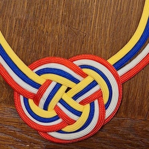 Könnte beinhalten: Eine Halskette aus rot, weiß, blau und gelbem Band, das zu einem Knoten geflochten ist. Die Halskette hat einen silbernen Verschluss.