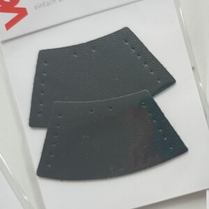 Può includere: Due toppe di pelle grigia con fori per la cucitura. Le toppe hanno la forma di un rettangolo curvo e sono etichettate "Kordelenden Lederimitat einfach annahen, fertig!"