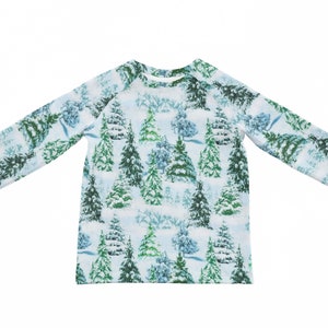 以下が含まれることがあります： 白い背景に、雪で覆われた緑と青の木が繰り返し描かれた長袖シャツです。