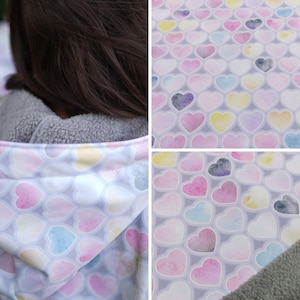 Può includere: Primo piano di una giacca con cappuccio con uno sfondo grigio chiaro e un motivo di cuori ad acquerello in tonalità di rosa, blu, giallo e viola. La giacca è foderata con un morbido pile grigio.