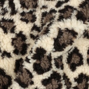 Könnte beinhalten: Ein plüschiger, strukturierter Stoff mit einem sich wiederholenden Leopardenmuster. Das Material hat einen dicken, flauschigen Flor mit einer Farbpalette aus cremefarbenen Grundtönen, die durch dunkelbraune und anthrazitfarbene Flecken akzentuiert sind.