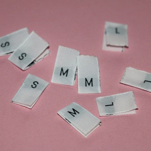 May include: White fabric tags with the letters S, M, and L printed on them. The tags are arranged on a pink surface.