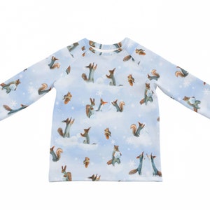 以下が含まれることがあります： 雪の中で遊ぶリスの模様がついた、薄い青色の長袖シャツ。リスは茶色と白で、雪は青みがかった白です。