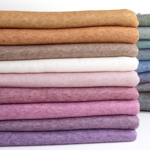 Könnte beinhalten: Ein Stapel gefalteten Stoffes in verschiedenen Brauntönen, Rosa, Lila und Weiß. Der Stoff ist weich und texturiert und scheint eine Art Strickware zu sein.