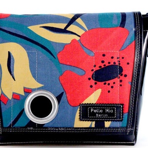 Könnte beinhalten: Eine schwarze Umhängetasche mit einem Blumendruck mit roten, gelben und blauen Blumen. Die Tasche hat einen schwarzen Lederriemen und einen Metallring an der Vorderseite. Die Tasche hat ein Etikett mit der Aufschrift "Pelle Mia Berlin".