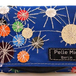 Könnte beinhalten: Eine blaue Geldbörse mit einem einzigartigen Design. Sie verfügt über farbenfrohe kreisförmige Akzente und weiß genähte Sternmuster. Die Geldbörse trägt den Text "Pelle Mia Berlin" auf einem schwarzen Etikett.