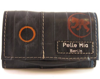 May include: A black wallet made from recycled inner tubes with white stitching. The wallet has a circular orange design, a red and orange graphic of a monkey, and a black label that reads "Pelle Mia Berlin".