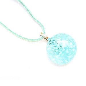Peut inclure: Un pendentif rond bleu clair avec des mouchetures bleu foncé est suspendu à un collier en cordon vert clair. Le pendentif est fixé à un fermoir de couleur argentée. Le collier est présenté sur fond blanc.