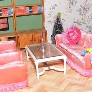 Può includere: Un set da salotto con un divano in velluto rosa, due poltrone in velluto rosa, un tavolino da caffè in vetro e un piccolo albero di Natale. Il divano ha un cuscino in crochet rosa e una coperta in crochet colorata.