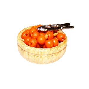 Op de afbeelding: Een miniatuur houten kom gevuld met oranje kralen, vergezeld van een zilverkleurige notenkraker. De kom heeft een lichtbruine, getextureerde uitstraling. De kralen zijn rond en gelijkmatig van formaat.