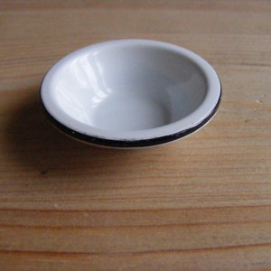 May include: A small, white enamel bowl with a black rim. The bowl is round and sits on a wooden surface. The bowl is empty and appears to be clean. The bowl is likely used for serving small portions of food or as a decorative item.