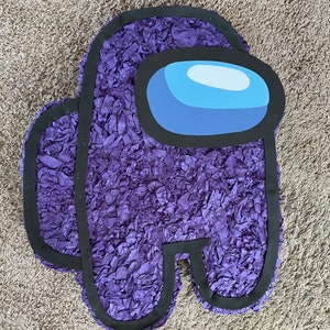 May include: A purple piñata in the shape of a video game character. The piñata is covered in ruffled purple paper and has a black outline. The character's visor is blue and white.