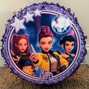 May include: Circular purple piñata featuring a central image of three animated women. The image is framed by a white border with white stars and a disco ball. The piñata is trimmed with purple fringe.