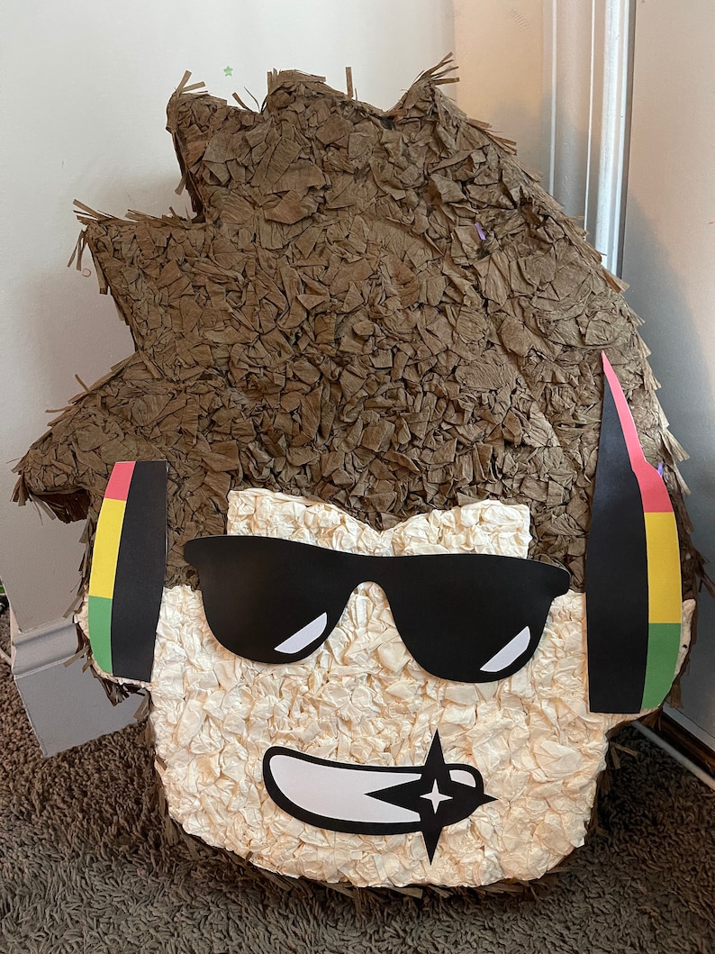 Op de afbeelding: Een bruine papier-mach&eacute; pi&ntilde;ata in de vorm van een persoon met een zonnebril en koptelefoon. De pi&ntilde;ata heeft een wit gezicht met een zwarte glimlach en een zwarte ster.