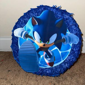 May include: Blue and white Sonic the Hedgehog pinata. The pinata features a cartoon image of Sonic running with his signature blue and white shoes.