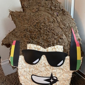 Op de afbeelding: Een bruine papier-mach&eacute; pi&ntilde;ata in de vorm van een persoon met een zonnebril en koptelefoon. De pi&ntilde;ata heeft een wit gezicht met een zwarte mond en een zwarte ster met een witte omtrek.