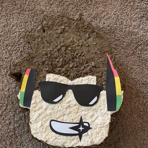 Op de afbeelding: Een bruine papier-mach&eacute; pi&ntilde;ata in de vorm van een cartoonfiguur met bruin haar, een zonnebril en een koptelefoon. De koptelefoon heeft rode, gele en groene strepen.