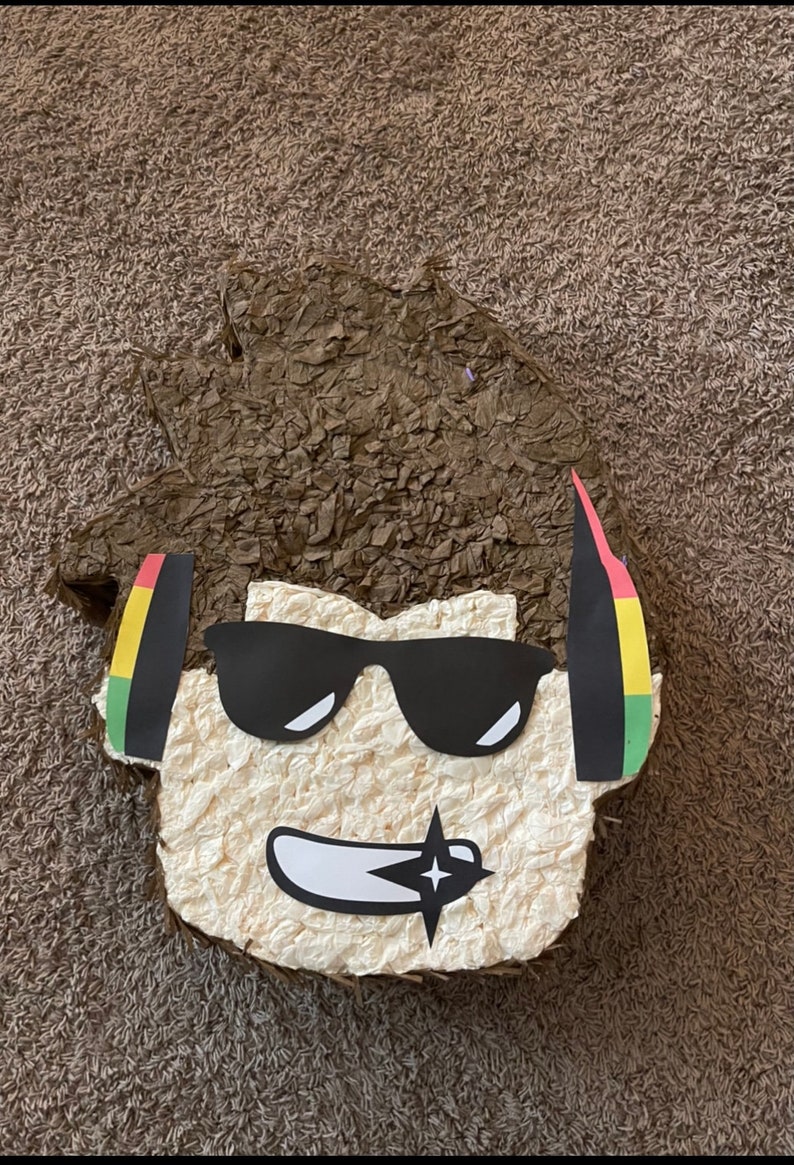Op de afbeelding: Een bruine papier-mach&eacute; pi&ntilde;ata in de vorm van een cartoonfiguur met bruin haar, een zonnebril en een koptelefoon. De koptelefoon heeft rode, gele en groene strepen.