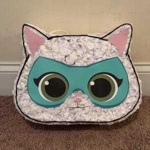 May include: White pinata shaped like a cat wearing a teal mask with large eyes. The pinata has pink ears and a white fluffy texture.