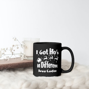 Op de afbeelding: Zwarte keramische mok met de tekst "I Got Ho's in Different Area Codes" in het wit. Het ontwerp bevat een kerstman-slee en rendieren. De mok staat op een wit oppervlak met een kaars en bloemen.