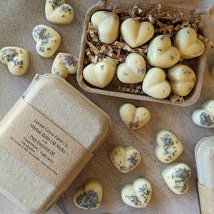 May include: Heart-shaped herbal bath oil melts in a cardboard box and scattered on a wooden surface. The melts are a creamy yellow color, some with visible herbs. The box is labeled "Locust Grove Farm Co. Herbal Bath Oil Melts" with peppermint and eucalyptus.