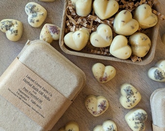 May include: Heart-shaped herbal bath oil melts in a cardboard box and scattered on a wooden surface. The melts are a creamy yellow colour, some with visible herbs. The box is labelled "Locust Grove Farm Co. Herbal Bath Oil Melts" with peppermint and eucalyptus.