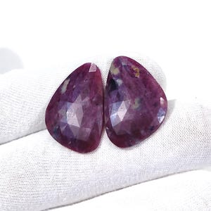 Könnte beinhalten: Zwei birnenförmige, facettierte Edelsteine in einem tiefen Magenta- und Violettton. Die Steine haben eine polierte, mehrfach facettierte Oberfläche, die das Licht reflektiert und natürliche Einschlüsse sowie Farbvariationen im Material zeigt.