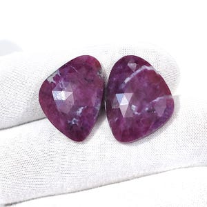 May include: Two loose, faceted, pear-shaped rubies with a deep magenta-red hue. The gemstones feature a polished, multi-faceted surface with visible internal white and dark inclusions, showcasing a natural, irregular, and organic gemstone texture.