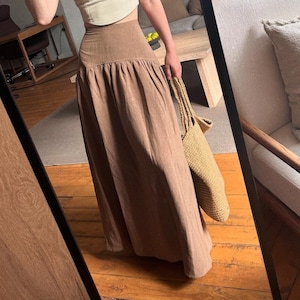 May include: A light cream-colored cropped corset-style top with thin straps paired with a high-waisted, floor-length brown maxi skirt featuring a wide waistband and a full, gathered silhouette, accompanied by a large woven straw tote bag.