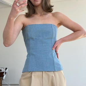 May include: A light blue denim strapless top with vertical seams. The top is paired with beige pleated pants. The model is holding a smartphone and wearing rings.