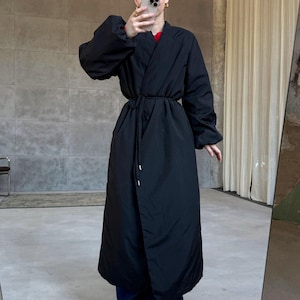 Peut inclure: Un long manteau noir léger de style matelassé, doté d'une fermeture croisée avec un fin cordon assorti à la taille. Ce manteau présente des manches longues et volumineuses avec des poignets élastiqués et une finition mate, arrivant aux chevilles.