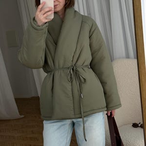 Puede incluir: Chaqueta acolchada verde oliva con cuello cruzado y cintura anudada. La chaqueta se lleva con vaqueros azul claro. La persona de la imagen sostiene un teléfono y se mira en un espejo.