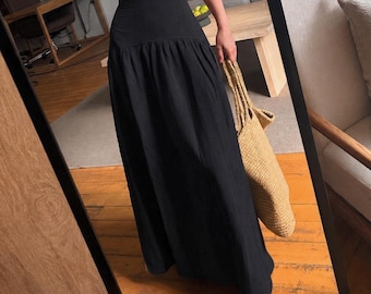 May include: A beige sleeveless crop top with thin straps and a straight neckline paired with a high-waisted black maxi skirt featuring a gathered waistline. A woven tan straw tote bag is held at the side, completing the ensemble.
