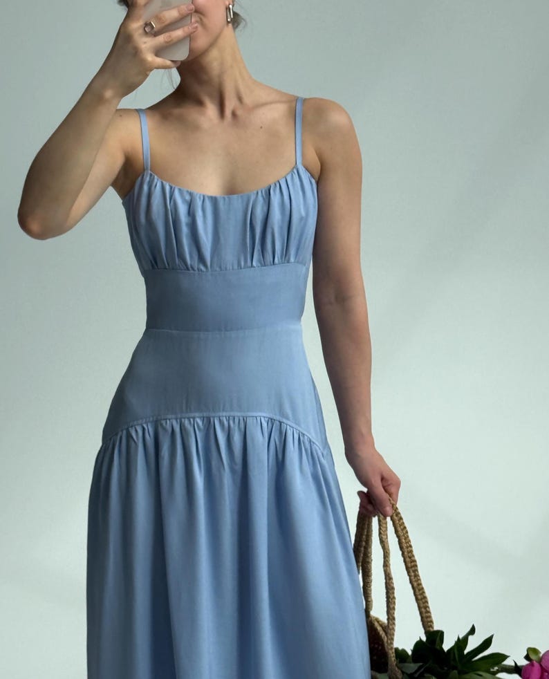 May include: A light blue midi dress featuring a scoop neckline, thin spaghetti straps, and a gathered bodice. The dress has a fitted waistline that transitions into a relaxed, flowing skirt with a horizontal seam detail below the waist.