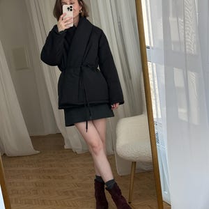 Puede incluir: Chaqueta acolchada negra con cintur&oacute;n envolvente, combinada con una falda verde oscuro y botas de ante burdeos. El conjunto se refleja en un gran espejo con marco dorado, creando un look elegante y moderno.