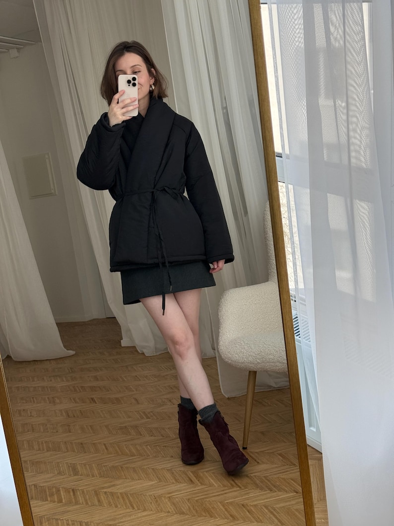 Puede incluir: Chaqueta acolchada negra con cintur&oacute;n envolvente, combinada con una falda verde oscuro y botas de ante burdeos. El conjunto se refleja en un gran espejo con marco dorado, creando un look elegante y moderno.