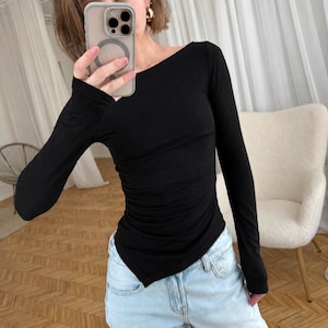 Könnte beinhalten: Ein schwarzes Langarmshirt mit weitem U-Boot-Ausschnitt und körpernaher Passform mit dezenter Raffung an den Seiten. Das Kleidungsstück hat einen spitz zulaufenden, asymmetrischen Saum, der seitlich leicht abfällt, kombiniert mit einer hellblauen Jeans.