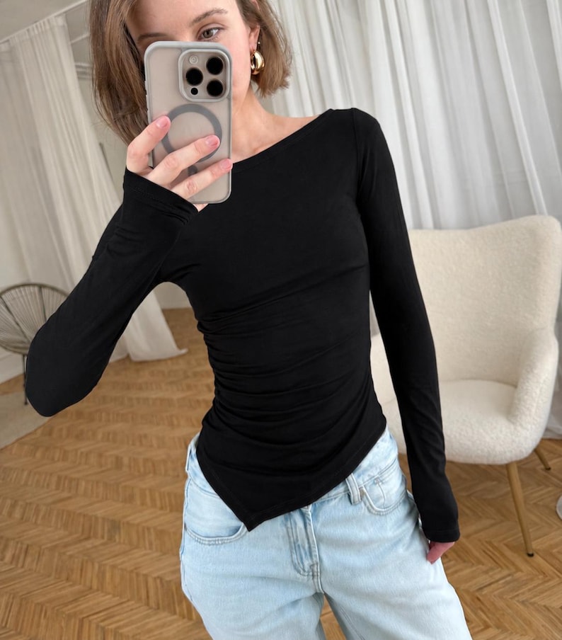 May include: A long-sleeved black top featuring a wide boat neckline and a fitted silhouette with subtle side ruching detail. The garment has a pointed, asymmetrical hemline that extends slightly at the sides, paired with light blue denim jeans.