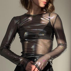 Può includere: Un top trasparente a maniche lunghe con finitura metallica iridescente. Il top ha uno scollo tondo e viene indossato sopra un capo intimo nero. Il tessuto riflette la luce, creando un effetto scintillante. Le maniche sono aderenti e lo stile generale è moderno ed elegante.